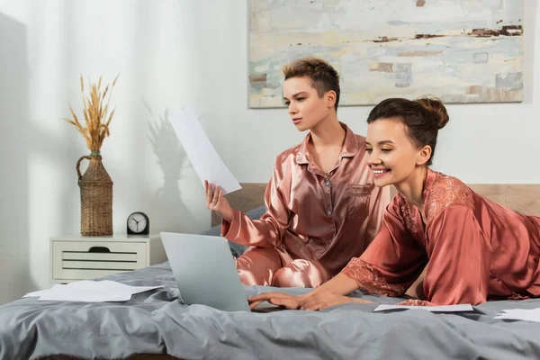 Persona grande feliz usando el ordenador portátil cerca de la pareja que trabaja con papeles en el dormitorio — Stock Photo