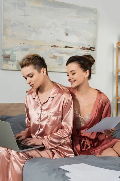 Happy pangender person holding document near partner typing on laptop in bedroom — Stock Photo