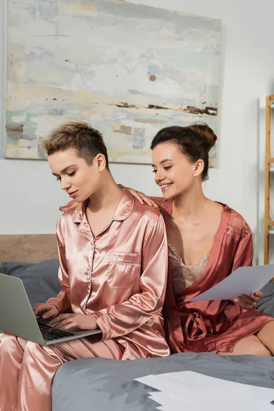 Happy pangender person holding document near partner typing on laptop in bedroom — стоковое фото