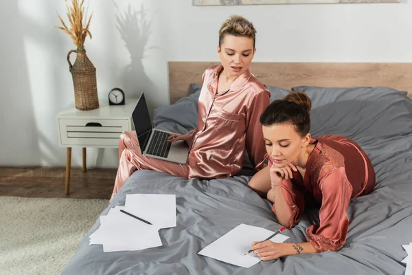 Young bigender person writing on paper near partner sitting on bed with laptop — стоковое фото