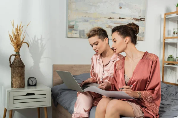 Pansexual partners working with papers and laptop in bedroom near wicker vase with spikelets - foto de stock