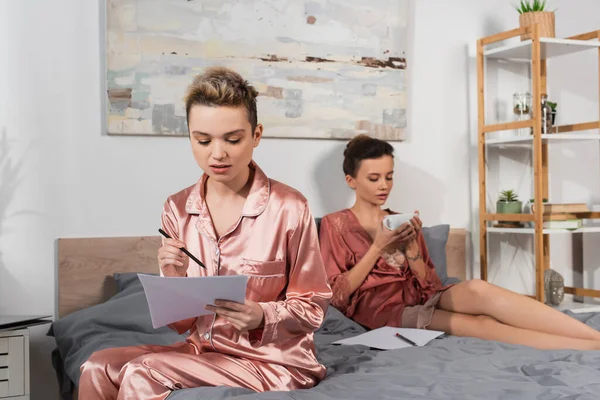 Pangender person in satin pajamas working with papers near partner drinking tea on bed — Stock Photo