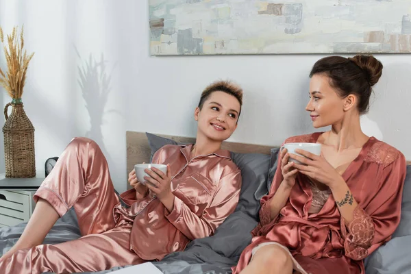 Smiling pangender couple in satin pajamas and robe drinking tea on bed - foto de stock