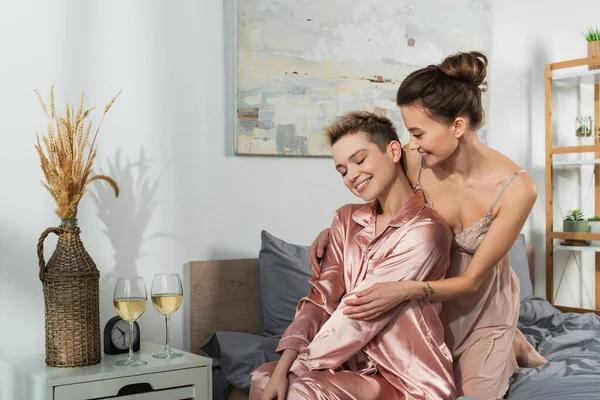 Smiling bigender person hugging young lover sitting in silk pajamas near glasses of white wine — Photo de stock