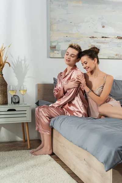 Young bigender person in silk pajamas sitting on bed near smiling partner and wine glasses — Stock Photo