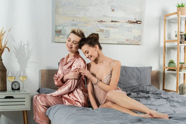 Young pansexual partners in sleepwear smiling near glasses of white wine in bedroom — стоковое фото