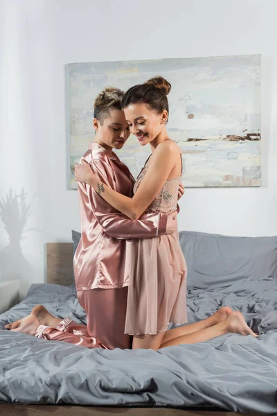 Young and happy pangender couple in sleepwear embracing on bed at home — Stock Photo