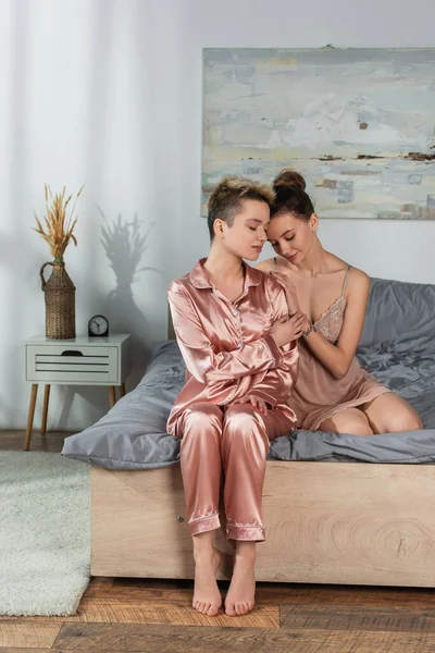 Young bigender person leaning on lover sitting on bed in satin pajamas — стоковое фото