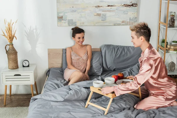 Young bigender person in satin pajamas holding tray with breakfast near happy lover — Photo de stock