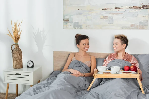 Young bigender lovers smiling at each other near tray with tea and chocolate toast bread - foto de stock