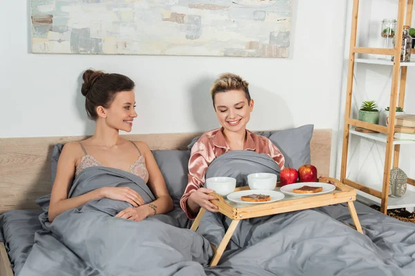 Jovens amantes pansexuais sorrindo perto da bandeja com café da manhã no quarto — Fotografia de Stock
