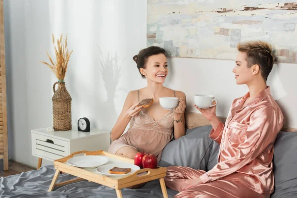 Cheerful pansexual lovers in sleepwear drinking tea with chocolate toast bread in bedroom - foto de stock