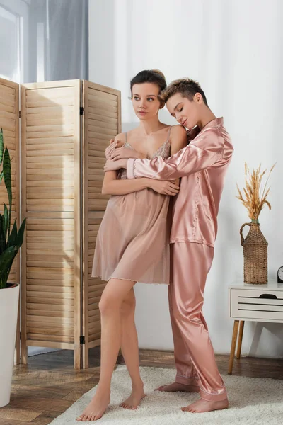 Full length of pansexual person in satin pajamas embracing partner looking away in bedroom — Stock Photo