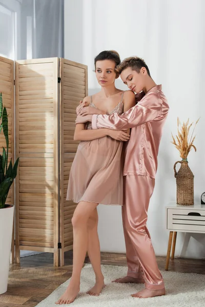 Full length of pansexual person in satin pajamas embracing partner looking away in bedroom — стоковое фото
