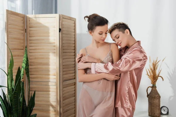 Bigender person in silk pajamas embracing young lover in bedroom — стоковое фото
