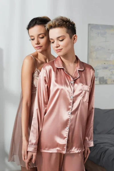 Bigender person in satin pajamas standing near young lover in bedroom — Fotografia de Stock