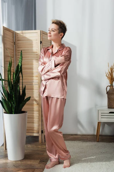 Full length of barefoot pangender person in satin pajamas standing with crossed arms near high flowerpot — стоковое фото