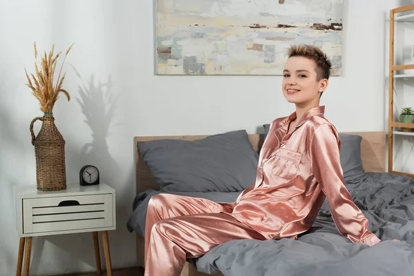 Smiling woman in silk pajamas sitting on bed near wicker vase with spikelets — Foto stock