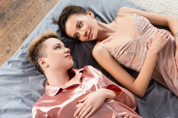 Top view of pansexual lovers in silk pajamas and nightdress lying on bed and looking at each other — стоковое фото