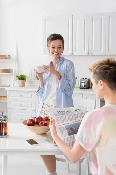 Cheerful pansexual person standing with tea cup near blurred partner reading newspaper during breakfast — Stock Photo
