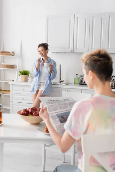 Blurred pangender person reading newspaper near happy partner with cup of tea — Photo de stock