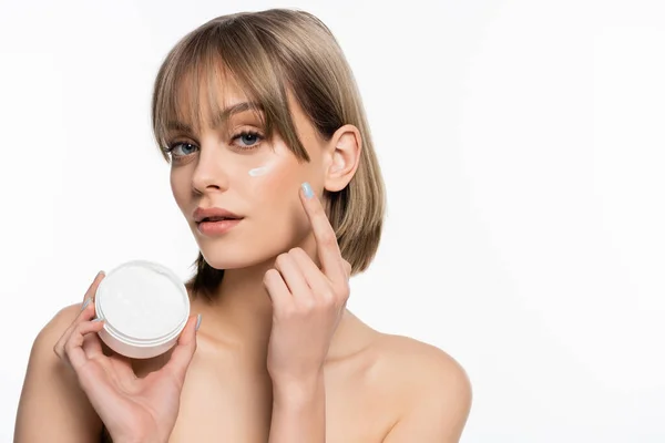 Young woman with bangs and cream on cheek holding container isolated on white — Stockfoto