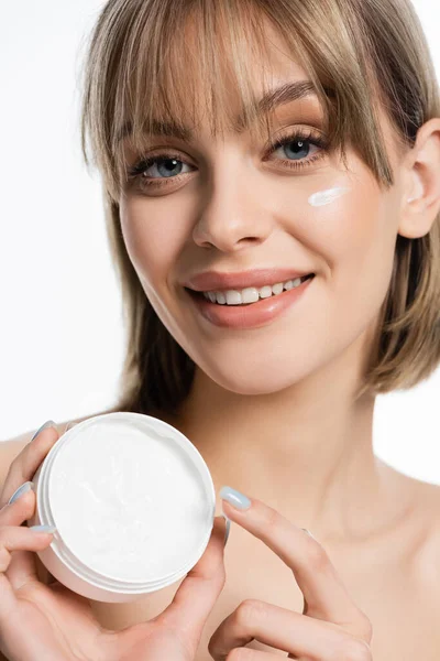 Joyful young woman with bangs and cream on cheek holding container isolated on white — Photo de stock