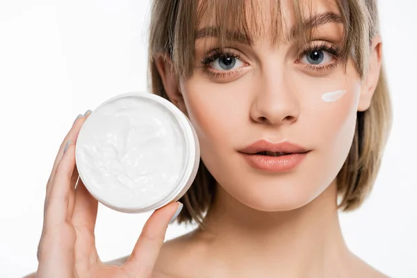 Young woman with bangs and cream on face holding container while looking at camera isolated on white — Fotografia de Stock