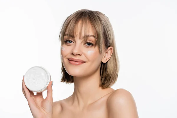 Smiling young woman with bare shoulders and cream on face holding container while looking at camera isolated on white — Photo de stock