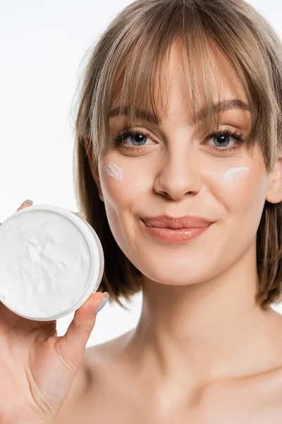 Smiling woman with bangs and cream on face holding container while looking at camera isolated on white — Stock Photo