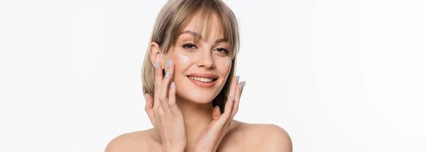 Happy woman with bangs applying cream on cheeks and looking at camera isolated on white, banner — Stock Photo