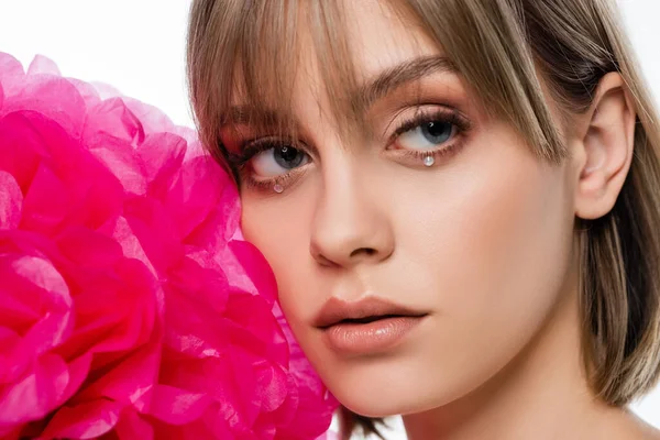 Pretty young woman with bangs and shiny rhinestones under blue eyes near pink flower isolated on white — Photo de stock