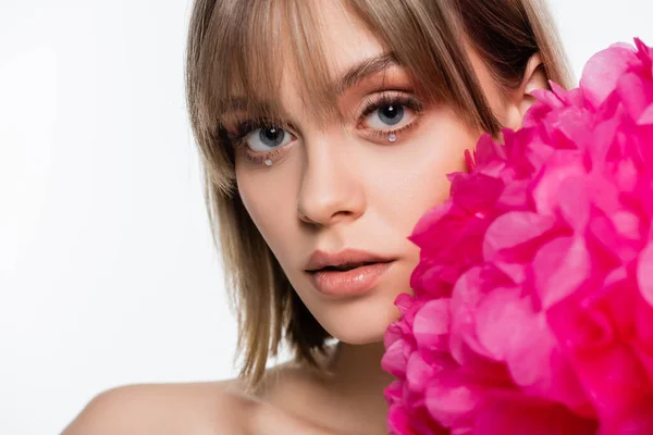 Mujer joven con flequillo y pedrería bajo ojos azules cerca de flor rosa aislada en blanco - foto de stock