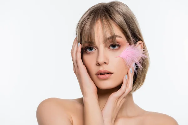 Sensual young woman with blue eyes and pink feather on cheek touching face isolated on white — стоковое фото