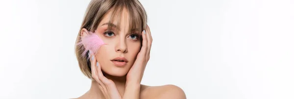 Jeune femme sensuelle aux yeux bleus et à la plume rose sur la joue touchant le visage isolé sur blanc, bannière — Photo de stock