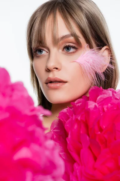 Jolie jeune femme avec plume sur la joue près de fleurs décoratives rose vif isolé sur blanc — Photo de stock