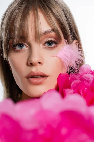 Pretty young woman with feather on cheek near blurred pink decorative flowers isolated on white — стоковое фото