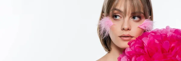 Jeune femme avec des éléments décoratifs dans le maquillage et des plumes roses sur les joues près de fleurs lumineuses isolées sur blanc, bannière — Photo de stock