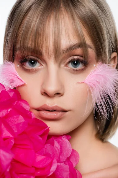 Gros plan de jeune femme avec des éléments décoratifs dans le maquillage et les plumes sur les joues près de fleur rose isolé sur blanc — Photo de stock