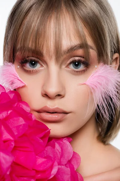 Close up of young woman with decorative elements in makeup and feathers on cheeks near pink flower isolated on white — Foto stock