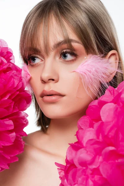 Mujer joven con elementos decorativos en maquillaje y plumas rosas en las mejillas cerca de flores aisladas en blanco - foto de stock