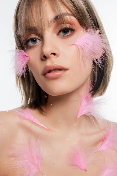 Primer plano de mujer joven con elementos decorativos y plumas de color rosa en la cara y el cuerpo aislados en blanco - foto de stock