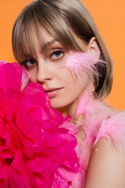 Young woman with decorative beads in makeup and feathers on cheeks near pink flower isolated on orange — Photo de stock