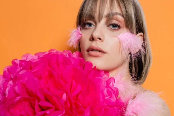 Pretty young woman with decorative beads in makeup and feathers on cheeks near pink flower isolated on orange — Photo de stock