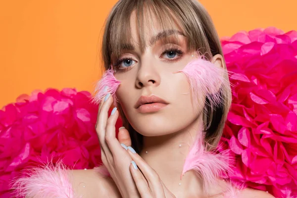 Blonde junge Frau mit dekorativen Elementen in Make-up und rosa Federn auf den Wangen in der Nähe von Blumen isoliert auf orange — Stockfoto