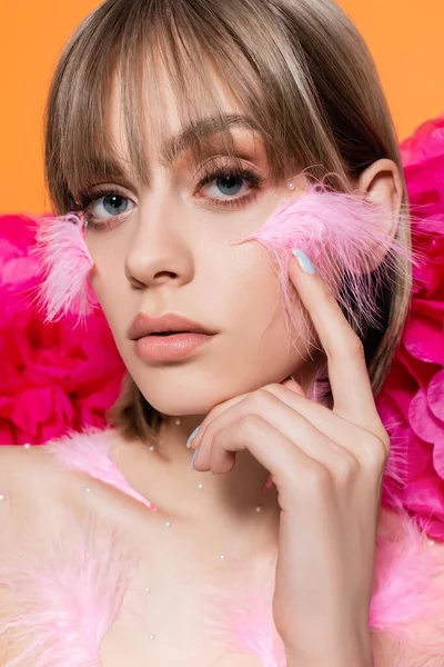 Jeune femme avec des éléments décoratifs en maquillage et plumes roses sur les joues posant près de fleurs isolées sur orange — Photo de stock