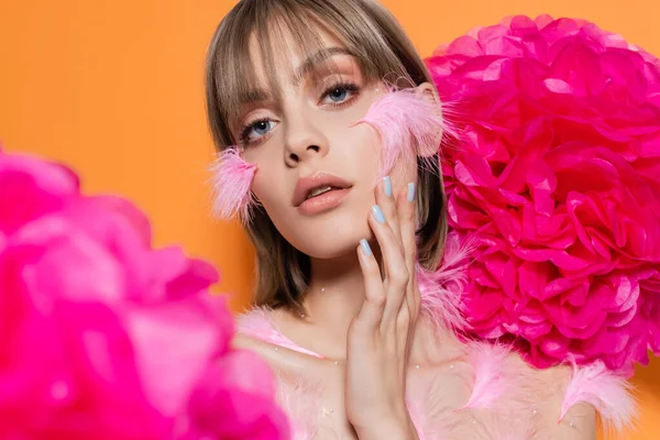 Mujer joven con elementos decorativos en maquillaje y plumas de color rosa en las mejillas cerca de flores aisladas en naranja - foto de stock