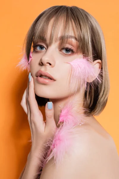 Mujer joven con cuentas decorativas en maquillaje y plumas de color rosa en la cara aislada en naranja - foto de stock