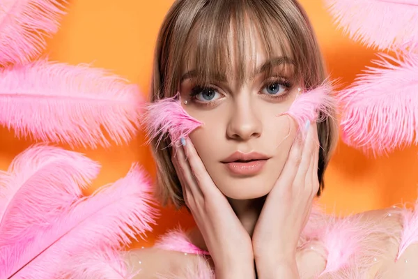 Junge Frau mit dekorativen Perlen im Make-up berührt Gesicht in der Nähe rosa Federn isoliert auf orange — Stockfoto