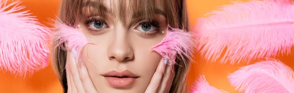 Mujer joven con cuentas decorativas en maquillaje tocando la cara cerca de plumas rosadas aisladas en naranja, pancarta - foto de stock